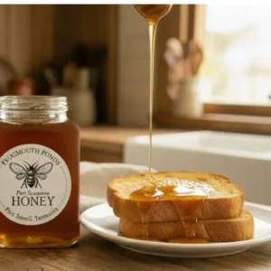 A jar of Frogmouth Ponds Bluegum Honey next to golden toasted bread with honey being drizzled on top in a rustic Tasmanian kitchen setting.