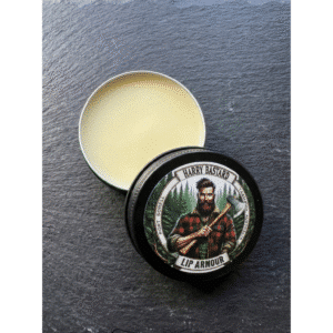 An open black tin of Harry Bastard Lip Armour showing the smooth, light-colored natural lip balm inside, resting on a dark textured slate surface with the branded lid leaning against it.