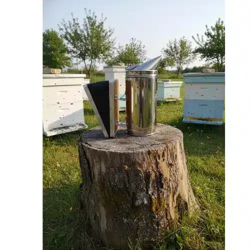 A stainless steel Beeco beekeeping smoker with black bellows sitting on a rustic wooden tree stump in a sunny outdoor apiary with white beehives in the background.